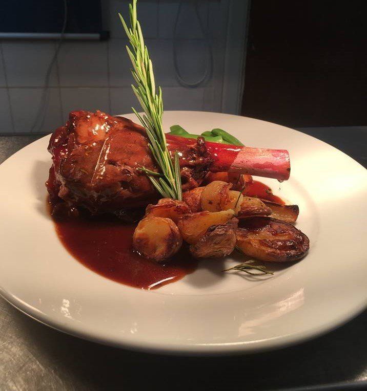 Braised meat shank, roasted potatoes, red sauce, rosemary garnish on a white plate.