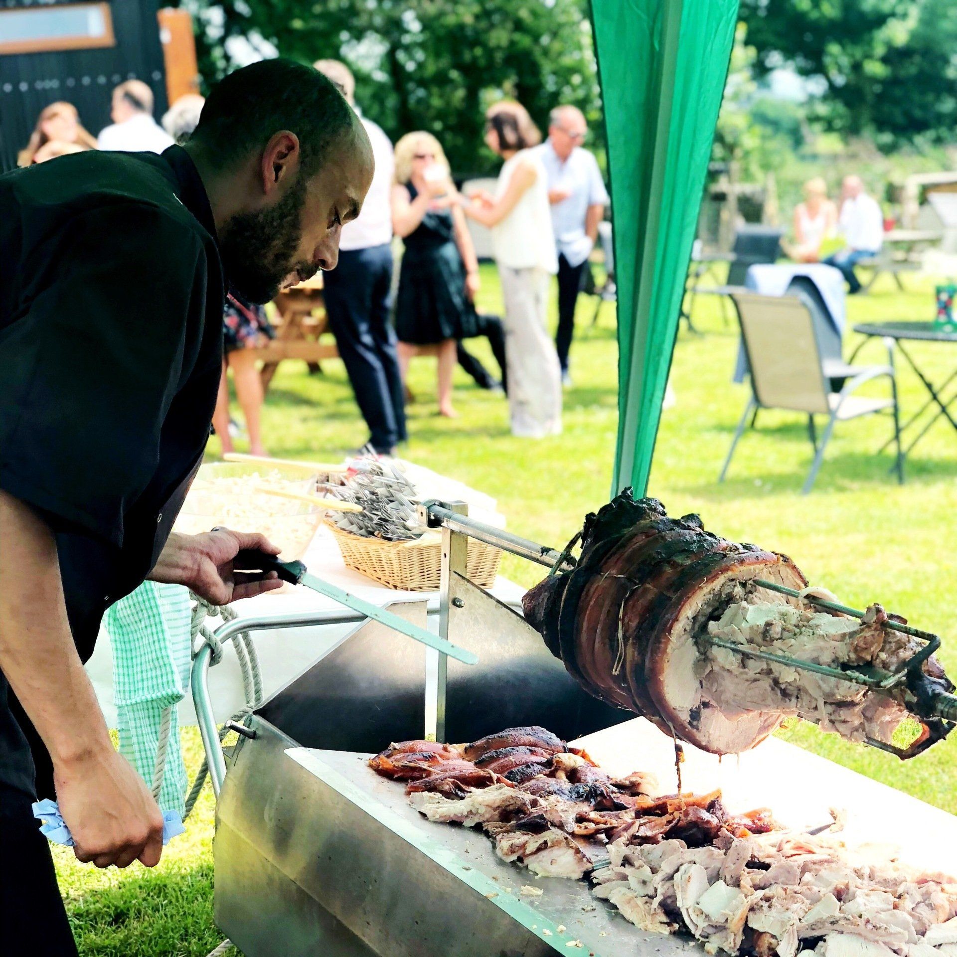 Chef carves roasted meat from a spit at an outdoor event; people in background.