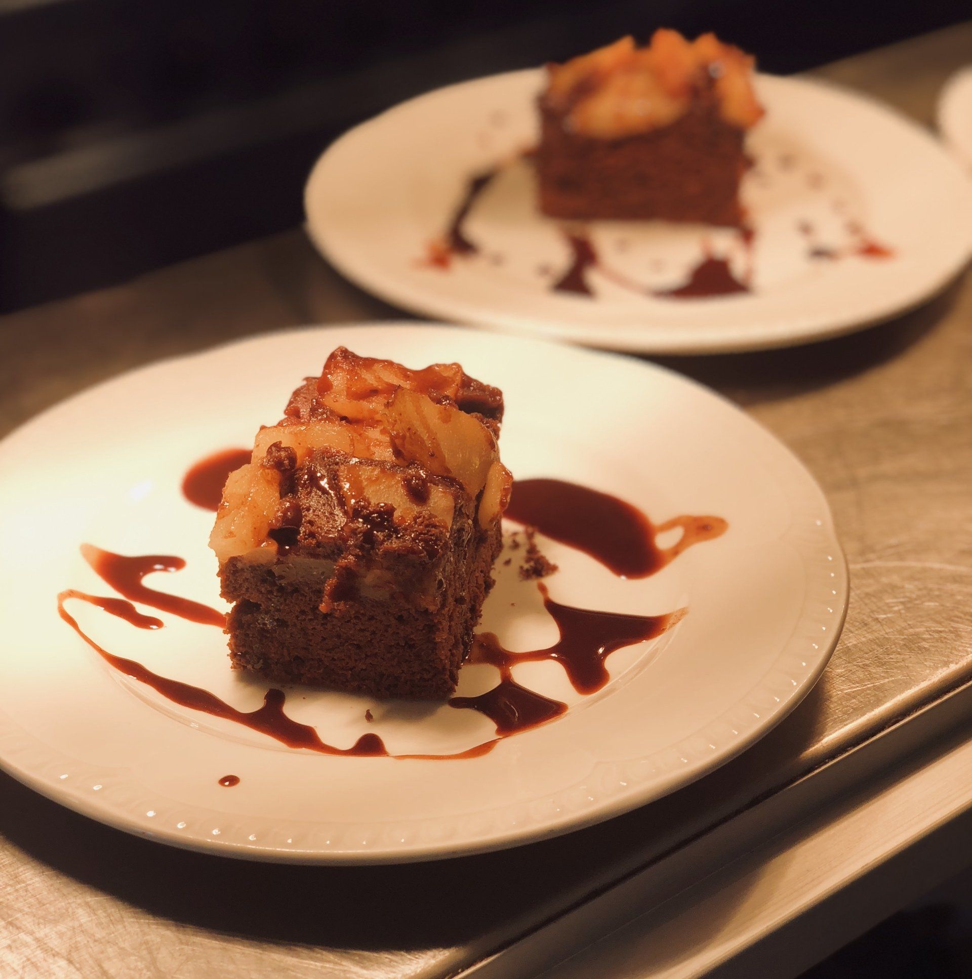 Two plates of chocolate cake topped with fruit and drizzled with sauce on a metallic surface.