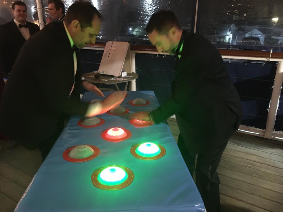 Two men in tuxedos playing a light-up game on a table. Setting appears to be an outdoor event.