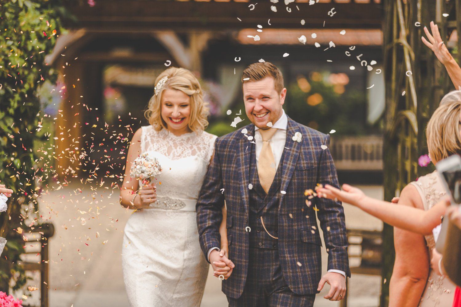 Newlyweds walking under confetti shower, smiling, holding hands. Outdoors, floral archway.