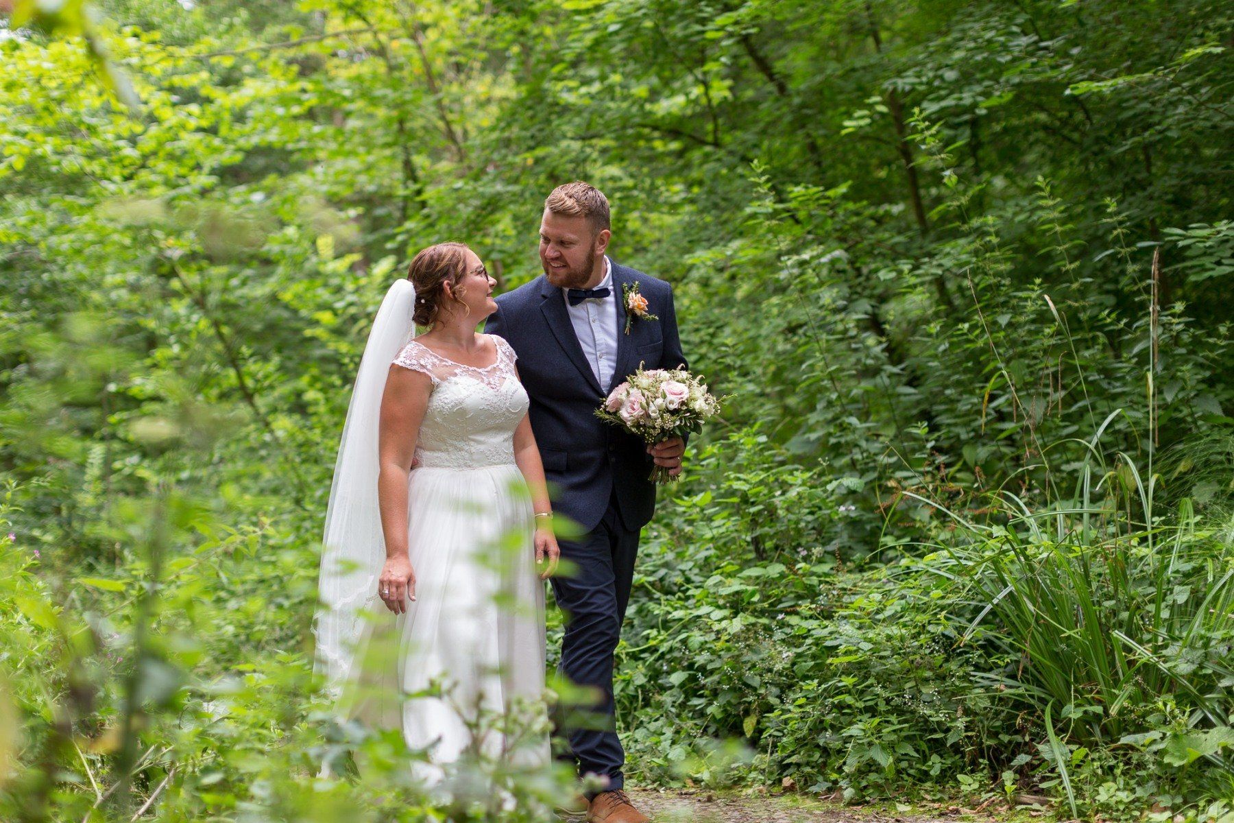 Bride and groom walking in a wooded area, smiling at each other. The groom holds a bouquet.