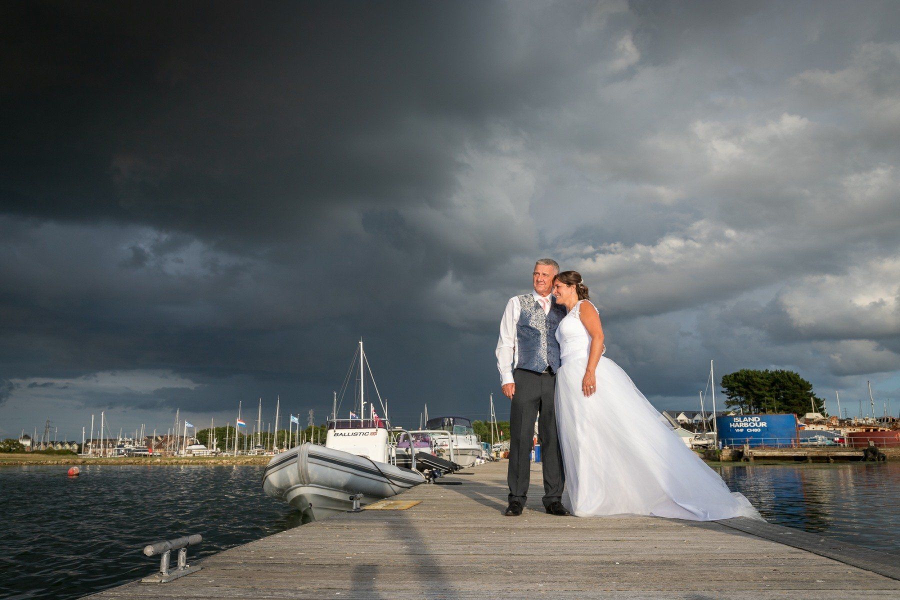 Bride and groom on a pier under a stormy sky near sailboats.