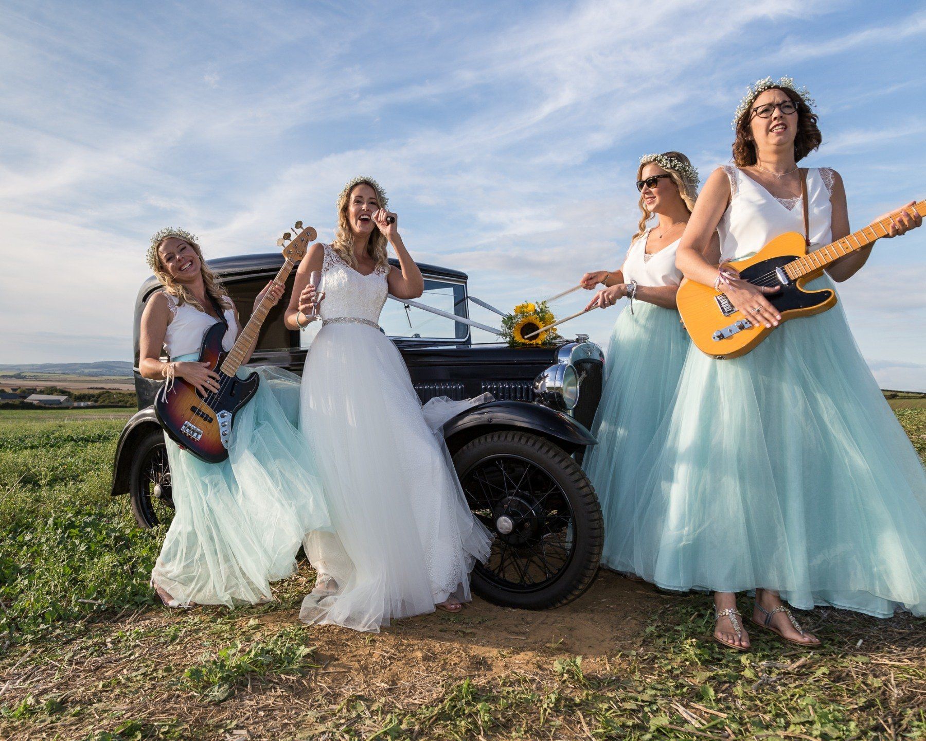 Bride and bridesmaids with guitars posing by vintage car in field. Sky is blue. All are smiling.
