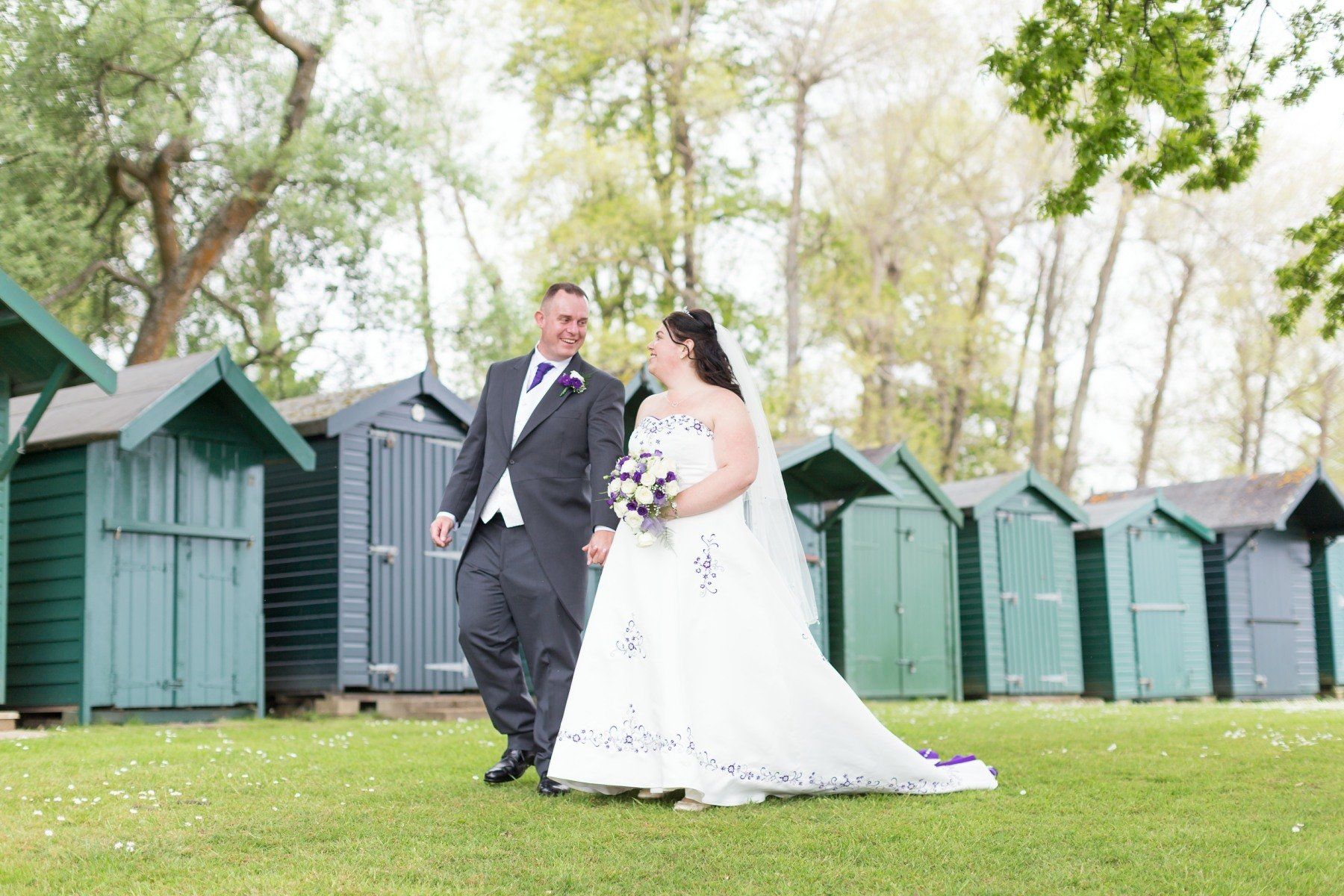 Bride and groom walking in a park, holding hands and smiling. Green sheds in the background.