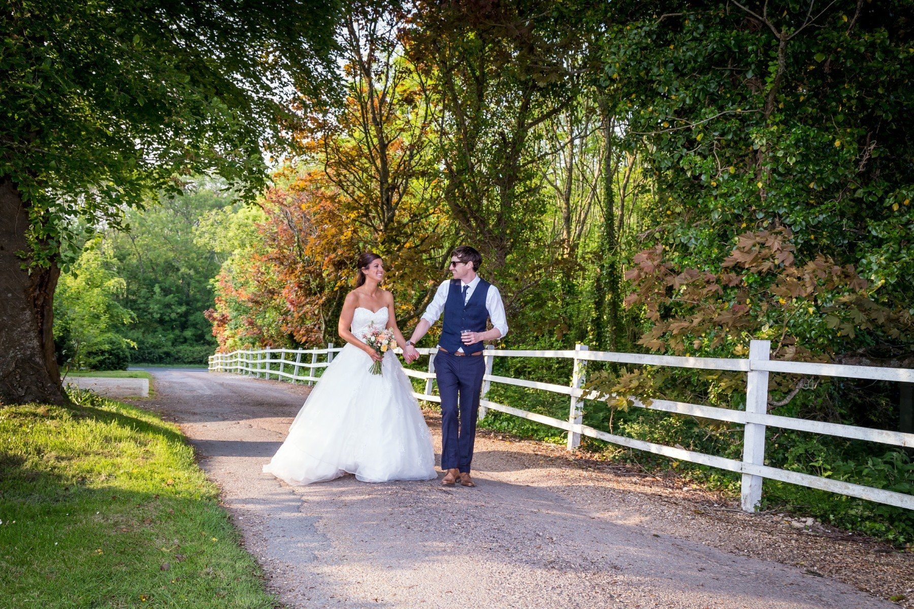 Bride and groom holding hands, walking on a gravel path near a white fence, surrounded by trees.