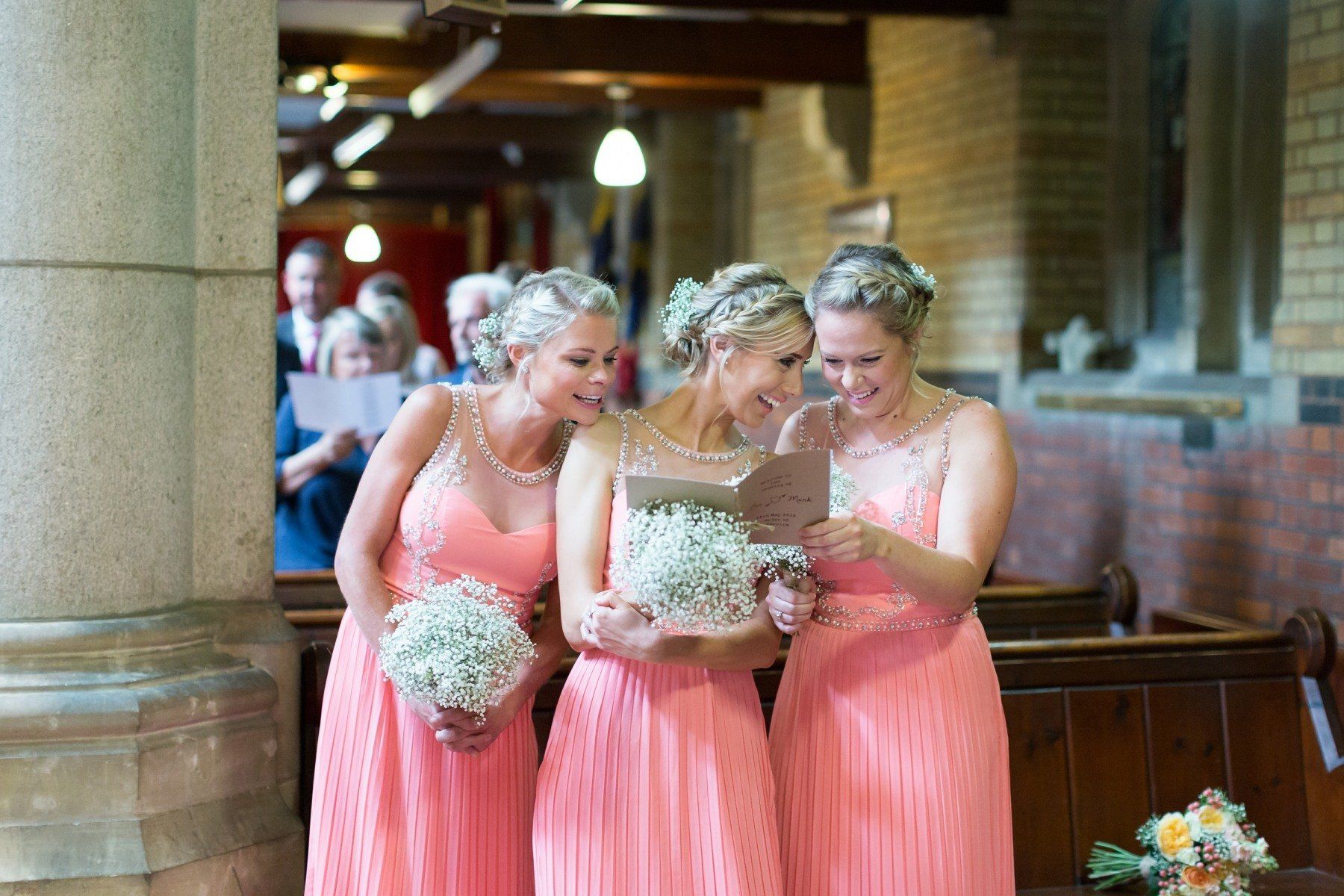 Three bridesmaids in coral dresses reading a card inside a church, holding bouquets.
