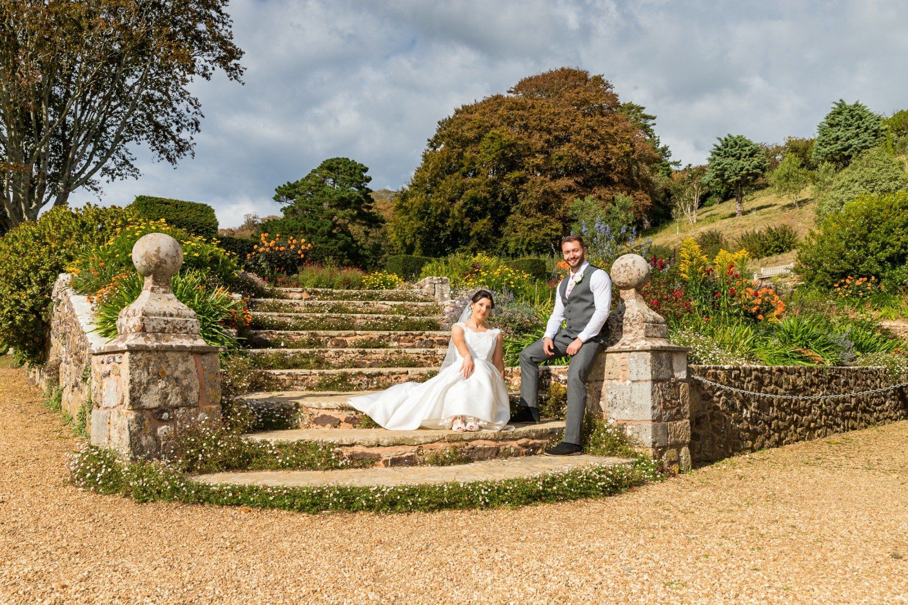 Wedding couple on stone steps in a garden; bride in white dress and veil, groom in vest, leaning.