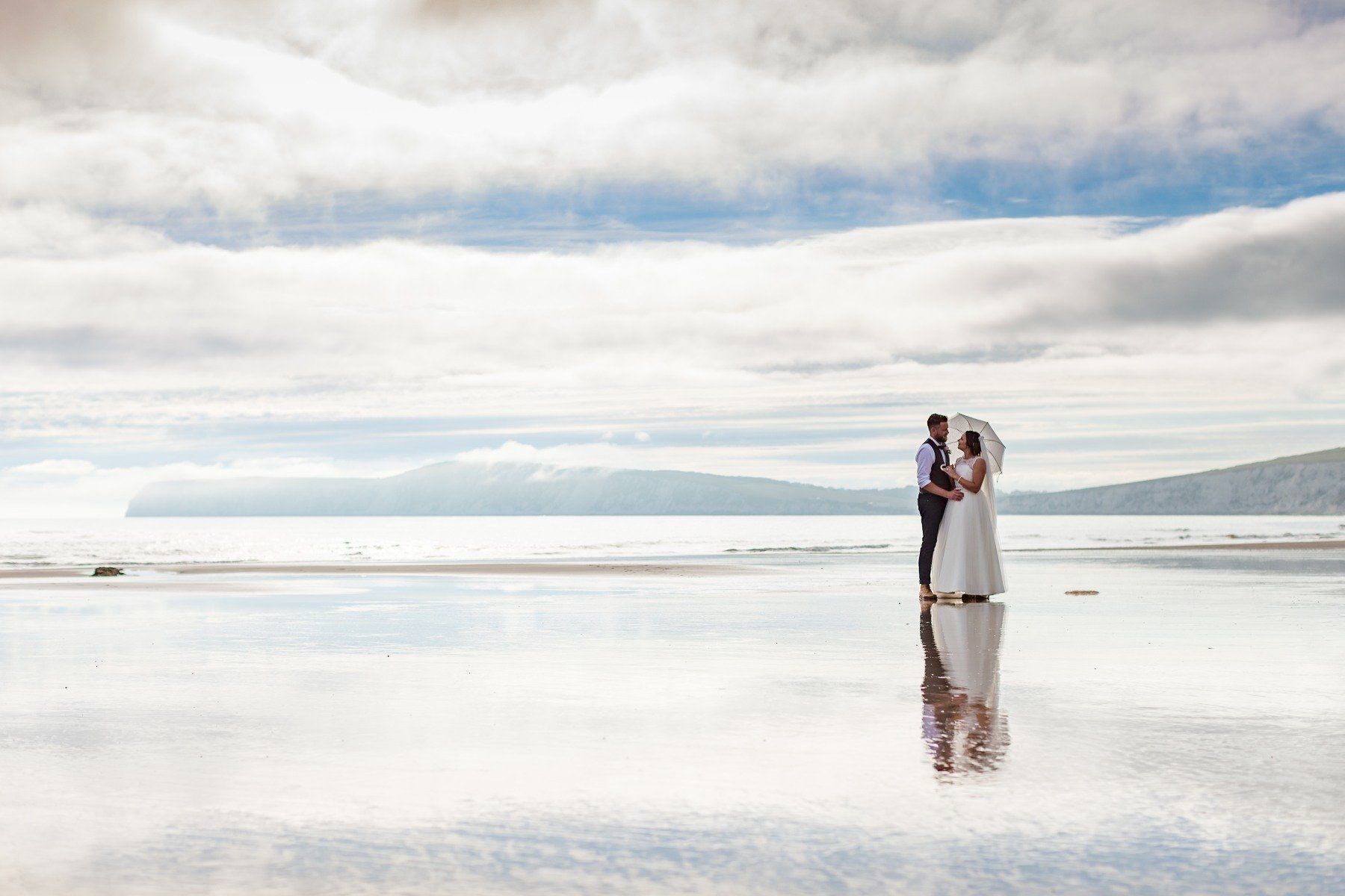 Couple in wedding attire on a reflective surface, mirroring sky and surroundings.