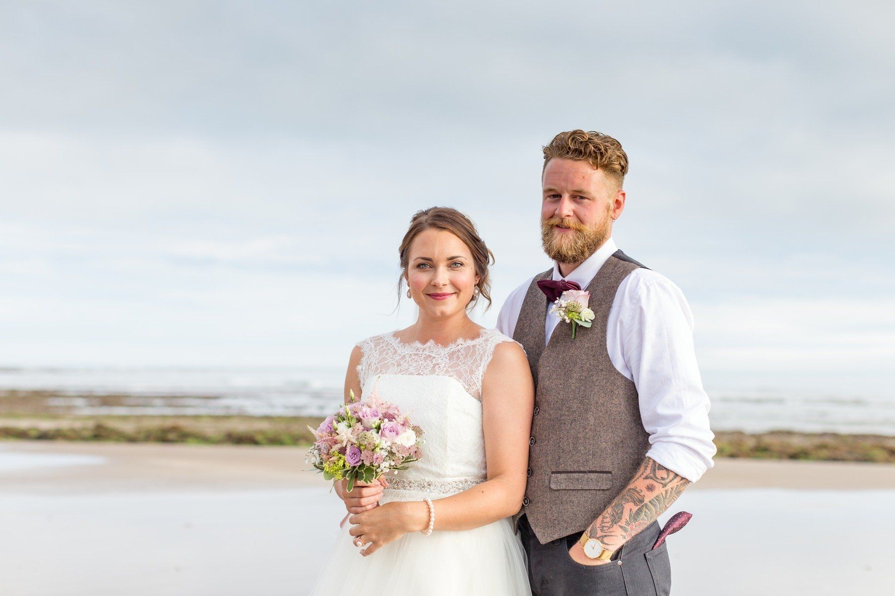Bride and groom stand on a beach. The woman holds a bouquet. The man has a beard and vest. Cloudy sky.