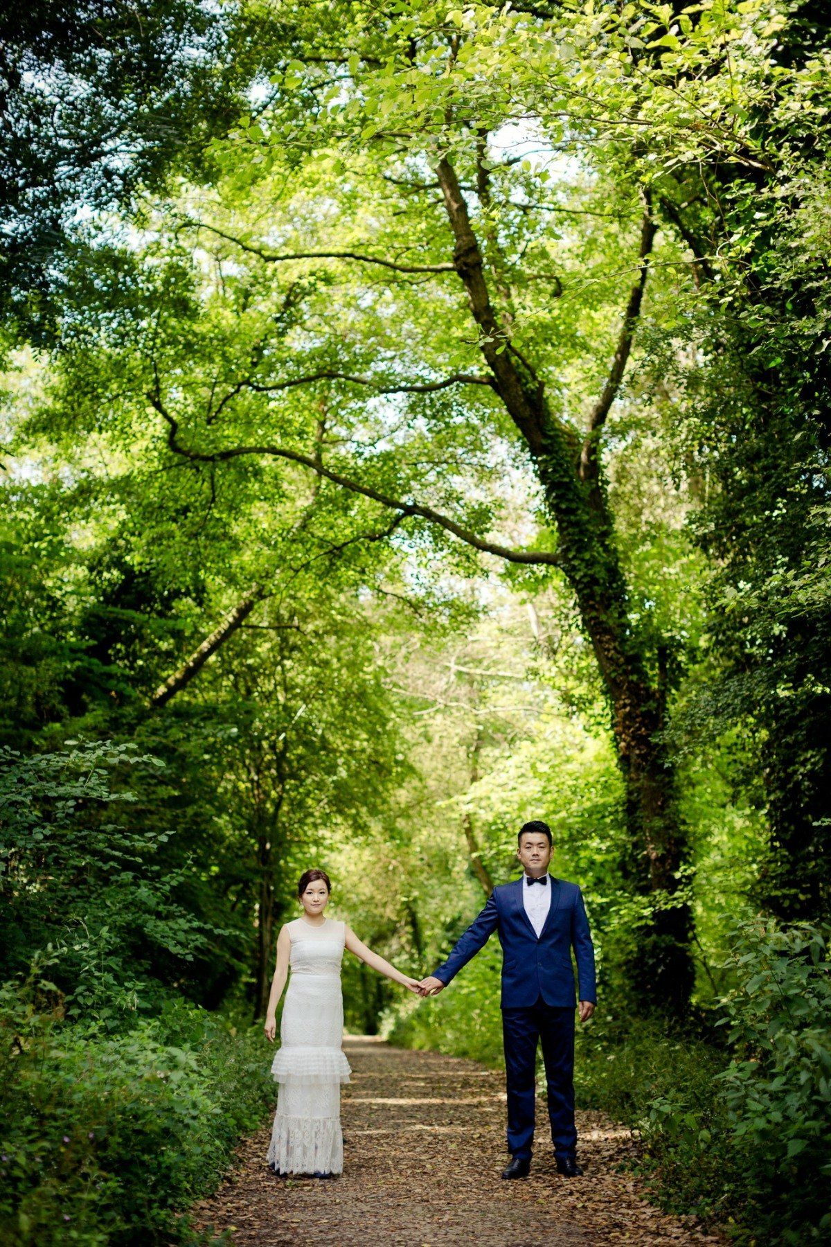 Couple in wedding attire holding hands on a path in a lush green forest.