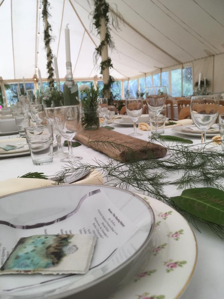Long table set for a formal event under a tent, with plates, glasses, and greenery.