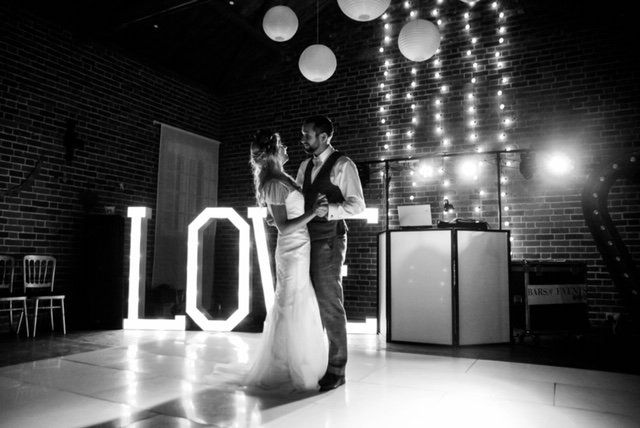Bride and groom dance beneath string lights, next to illuminated 