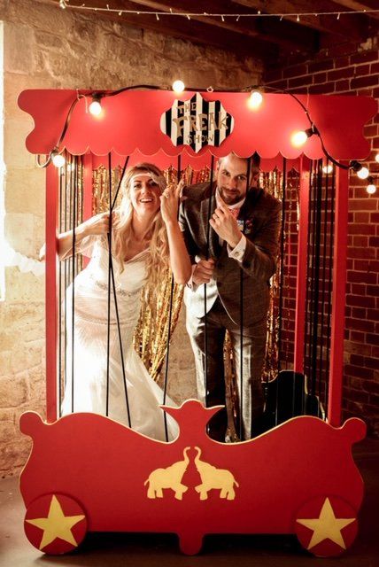 Wedding couple posing in a red circus-themed photo booth with string lights and a gold fringe backdrop.
