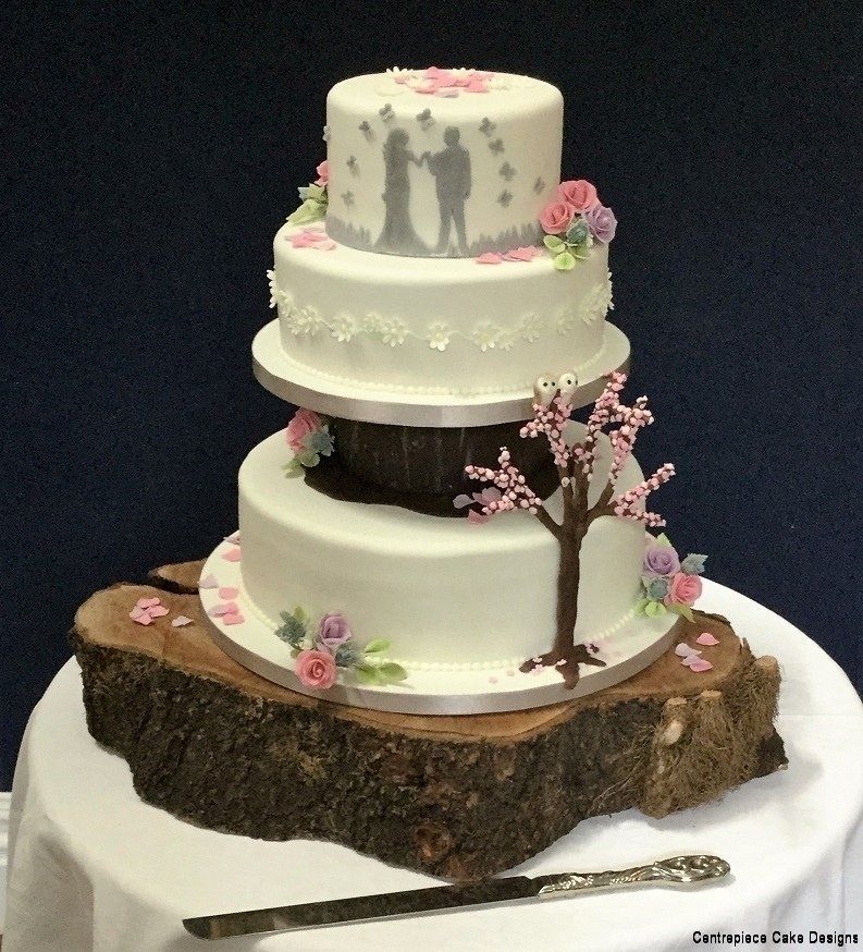 Three-tiered wedding cake with floral decorations, a tree design, and a bride and groom silhouette, on a wood slab.