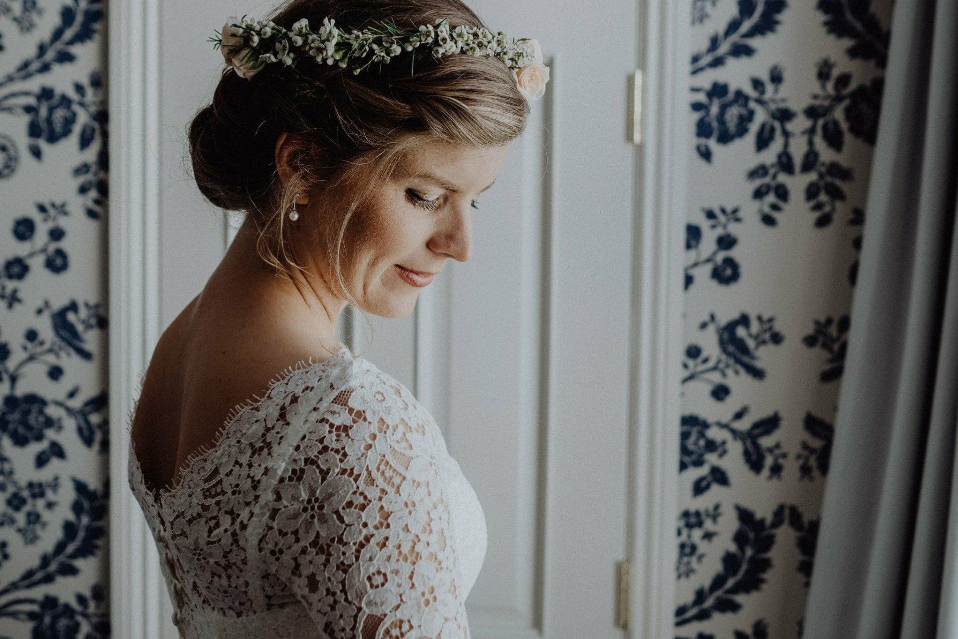 Bride in lace dress with flower crown, looking down. Interior with floral wallpaper and curtain.