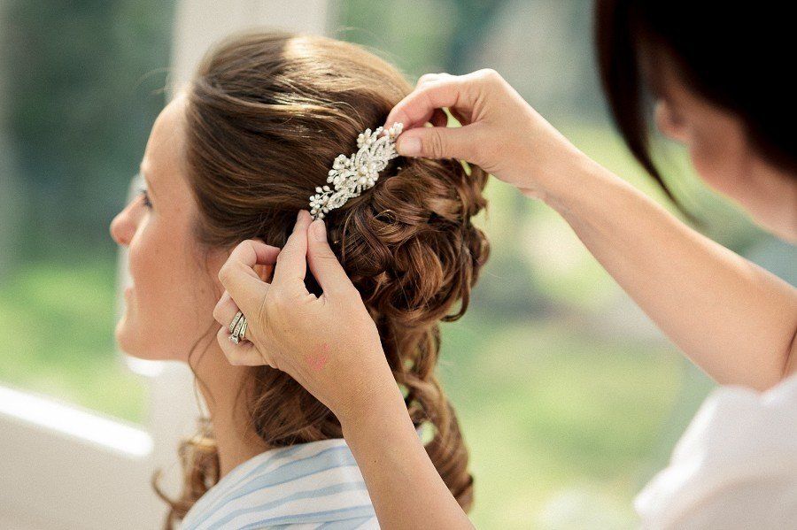 Woman's hair styled with decorative clip by another person's hands indoors, near a window.