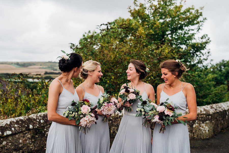 Four women in gray dresses holding bouquets, laughing together outdoors.