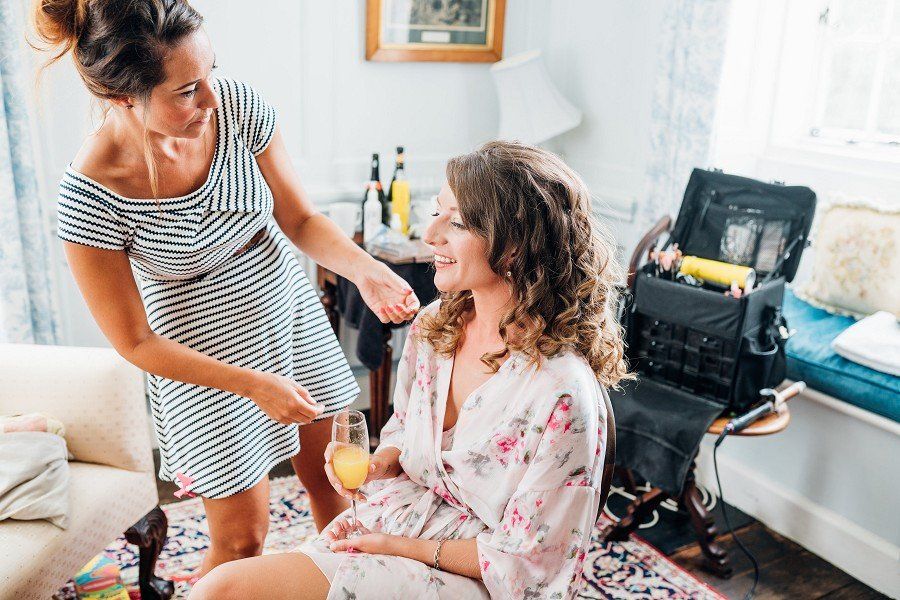 Woman in floral robe has hair styled; another woman styles it near a makeup kit indoors.