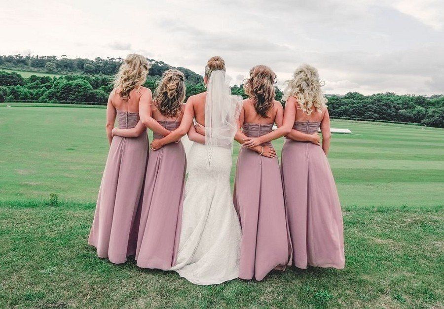 Bride and bridesmaids in mauve dresses, standing on a grassy hill, arms around each other.