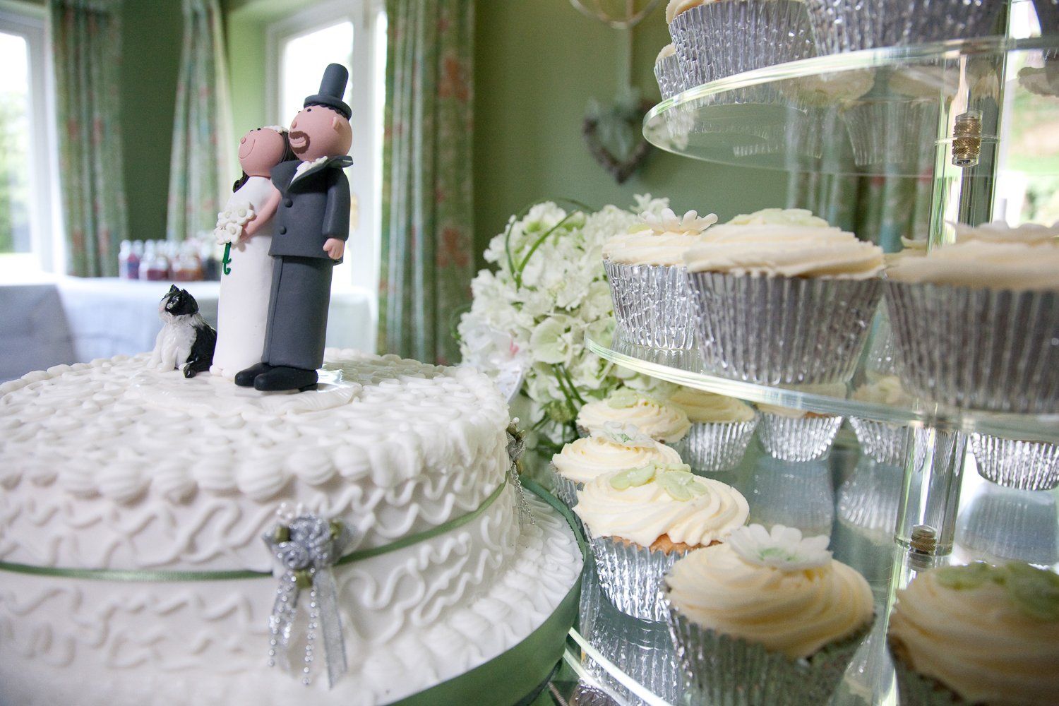 Wedding cake with bride and groom topper, cupcakes on tiered stand, and flowers in a light-filled room.