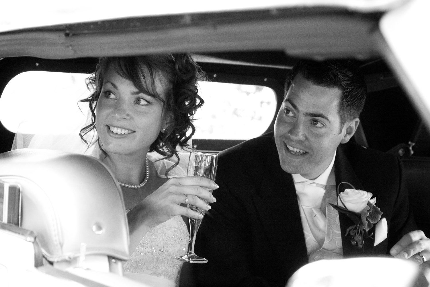 Newlyweds in a car, smiling, woman holding champagne, man wearing a suit with a boutonniere.