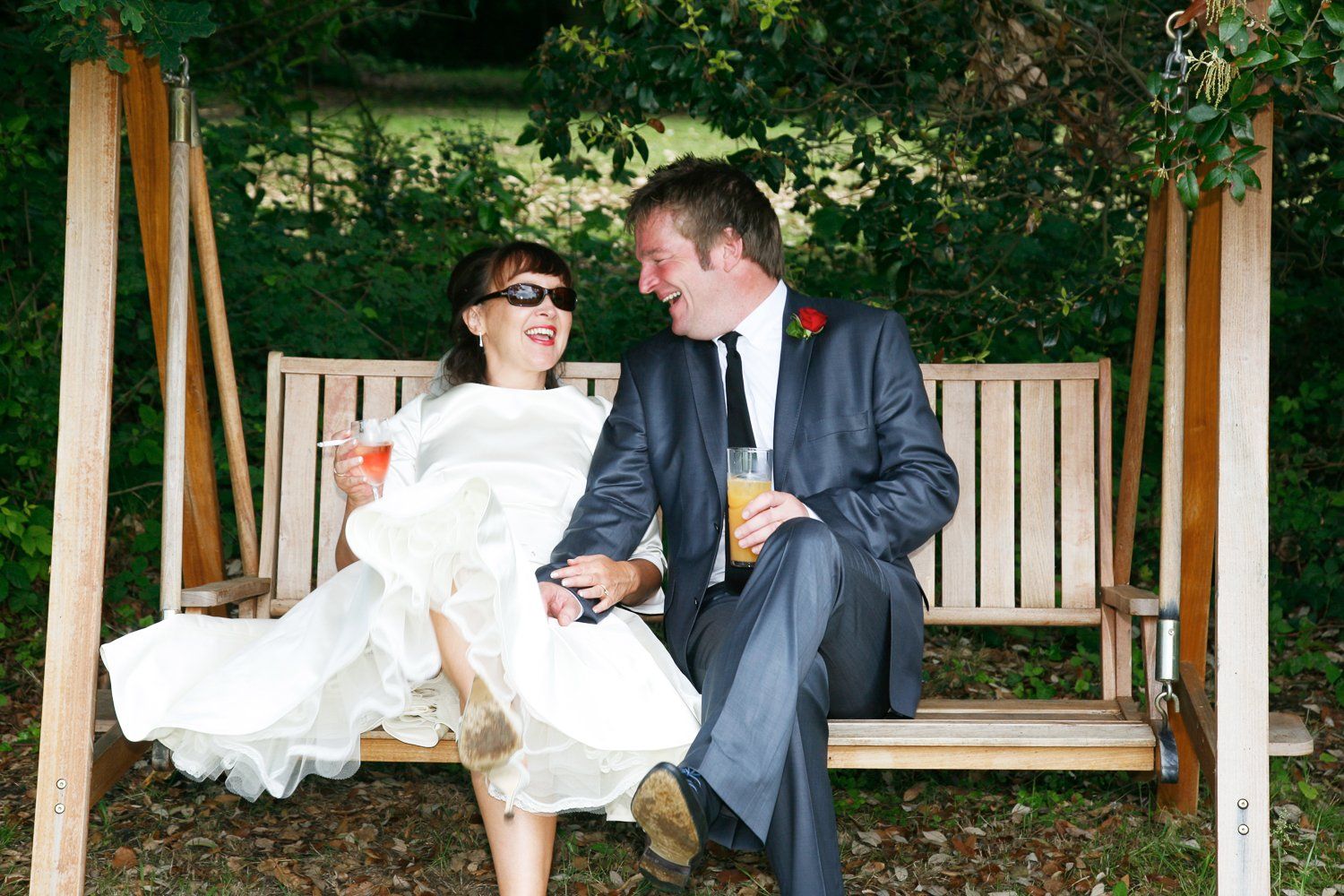 Newlyweds on a wooden swing, laughing and holding hands, outdoors. Bride in sunglasses, groom in suit.