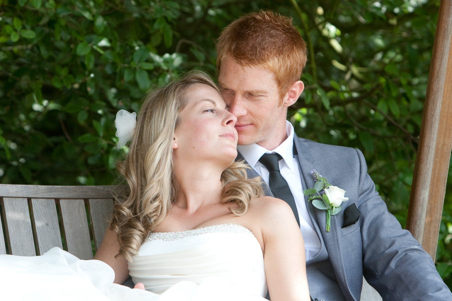 Wedding couple, light-haired woman and red-haired man, sit closely on a bench, looking at each other, outdoors.