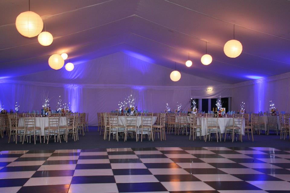 Event tent with checkered dance floor, round tables, chairs, and hanging globe lights with blue up lighting.