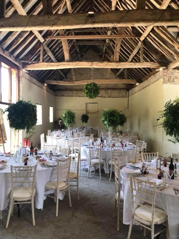 Barn interior set for a reception, with round tables, white tablecloths, and floral decorations. Wooden beams and hanging greenery.