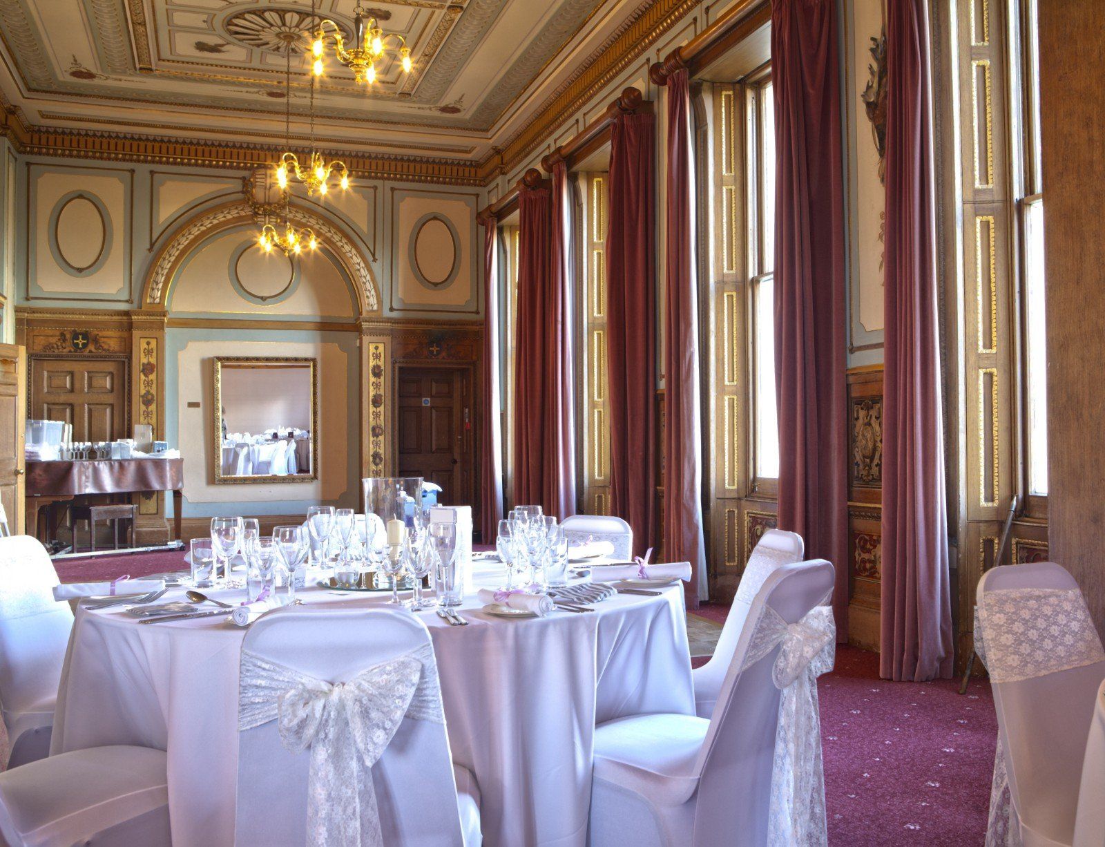 Elegant dining room with white-covered tables, set for a formal event. High ceilings, ornate details, and red curtains.