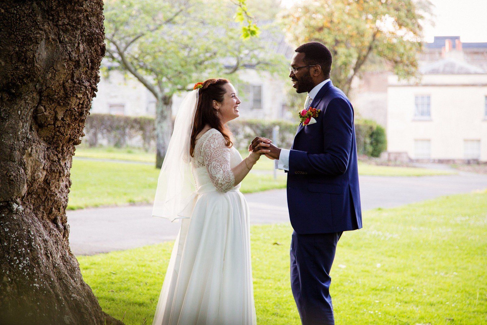 Couple holding hands, laughing, near a tree. She wears a white dress and veil, he wears a blue suit.