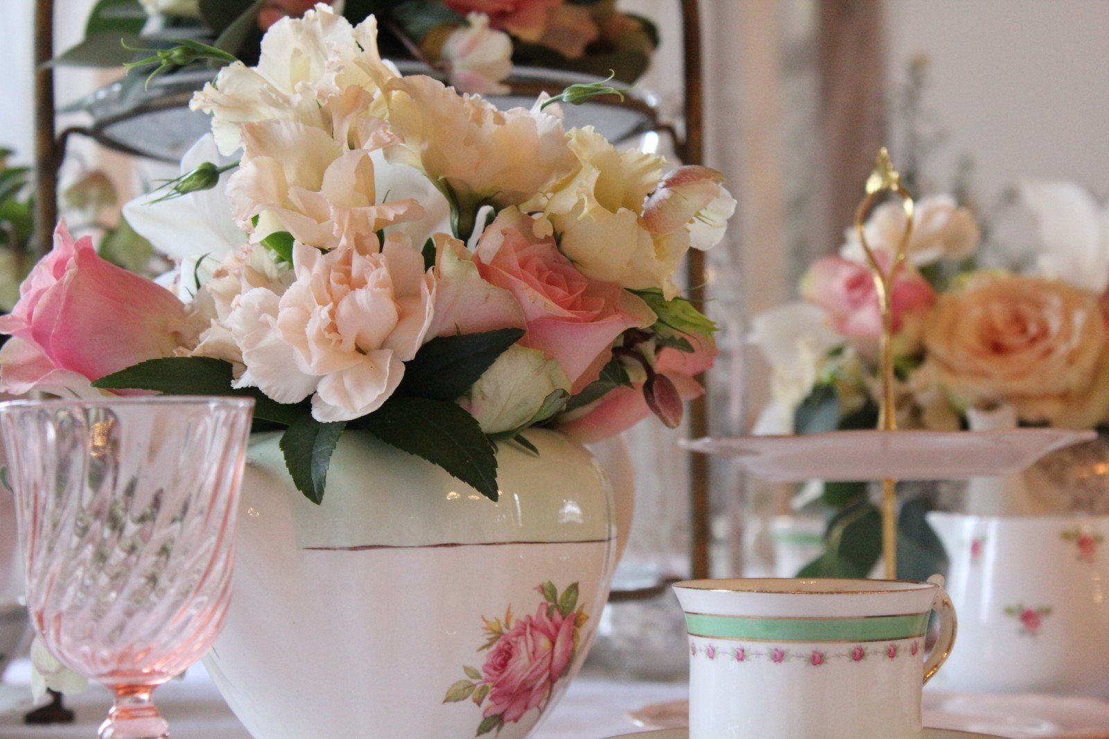 Floral arrangement in a decorated vase with teacups and a tiered stand, soft pink and cream colors, elegant setting.