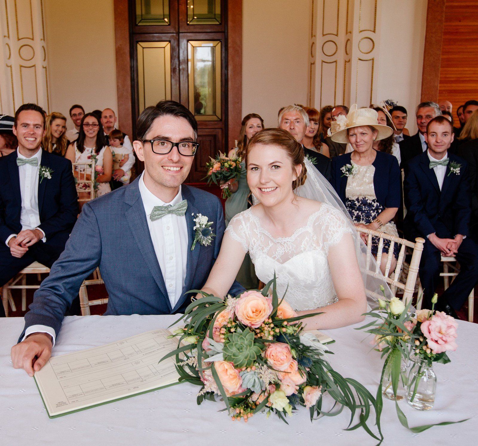 Couple signing marriage documents at a ceremony, surrounded by guests. Soft lighting, flowers, and smiles.