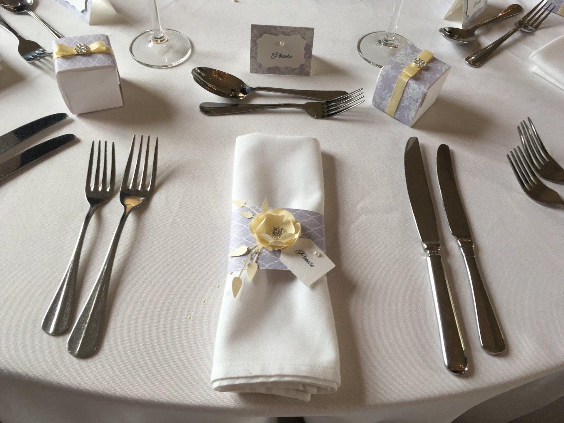 Table setting with white tablecloth, silverware, napkin with flower embellishment, and small gift boxes.