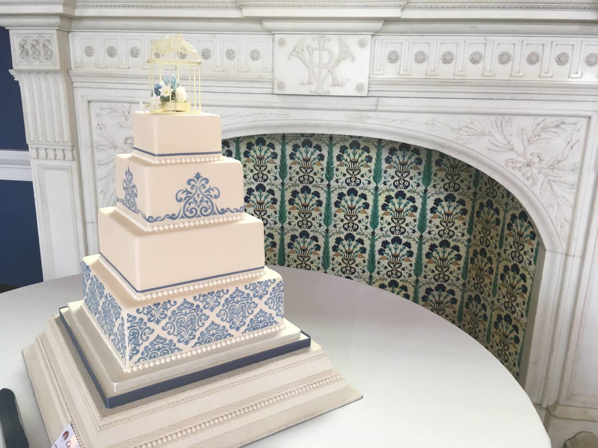 Five-tiered wedding cake with blue and white designs on a table, in front of a decorative fireplace.