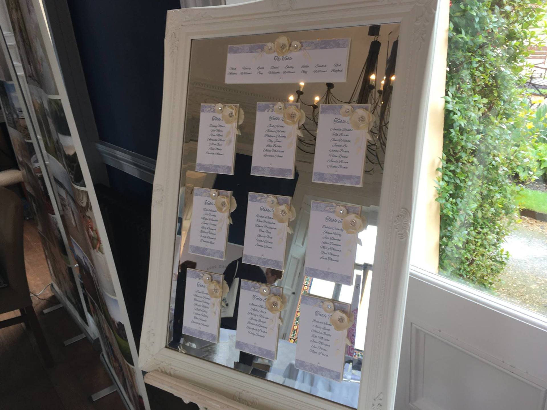 Mirrored seating chart with floral accents, lists names. In front of a window, white frame.