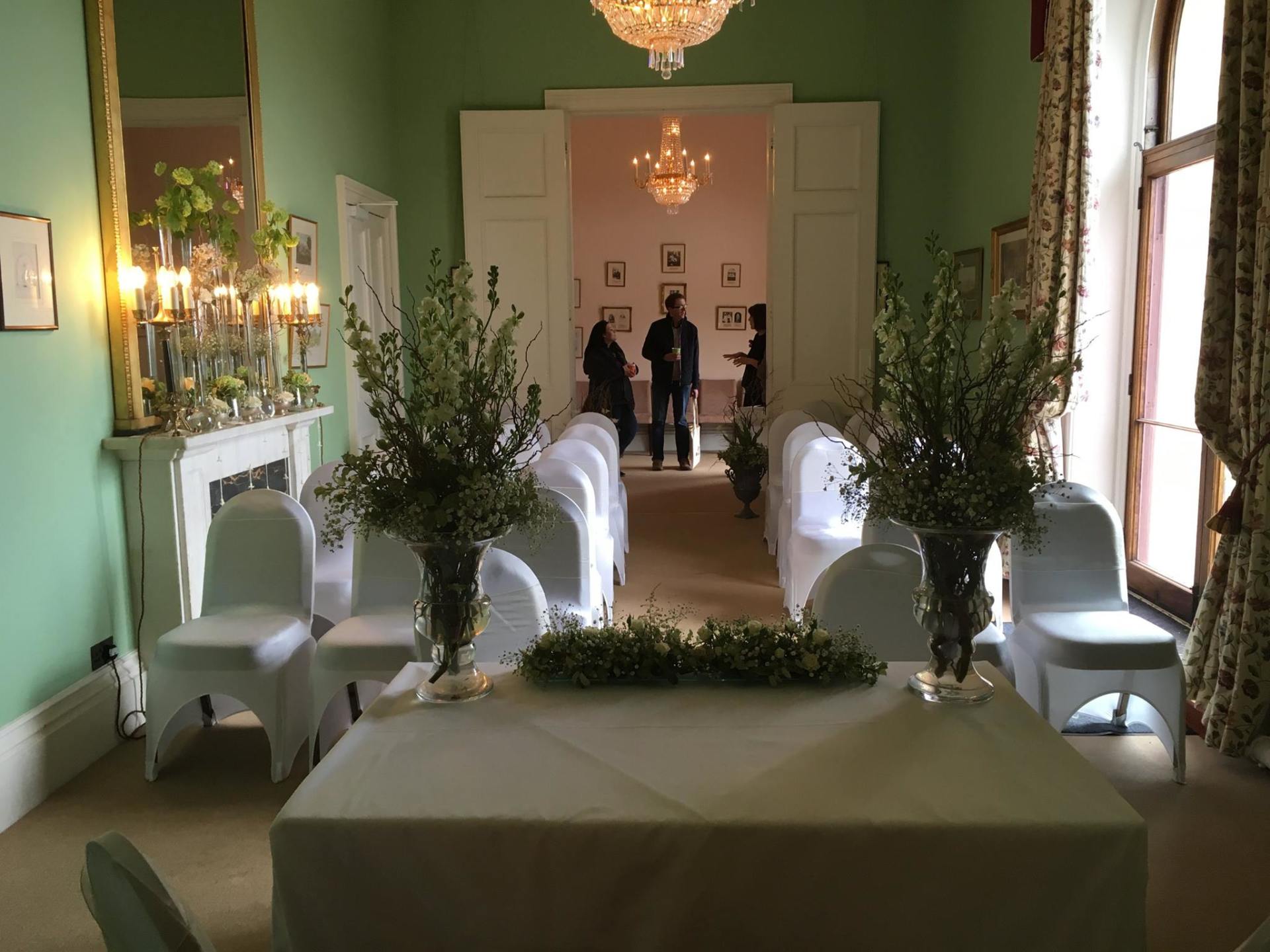 Wedding ceremony setup in a pale green room with floral arrangements and seated guests; the room has a chandelier and fireplace.