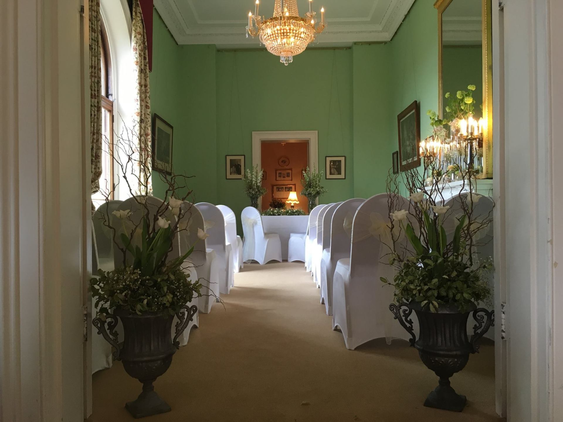 Wedding ceremony setup in a room with green walls and white-covered chairs. Urns with greenery flank the aisle.