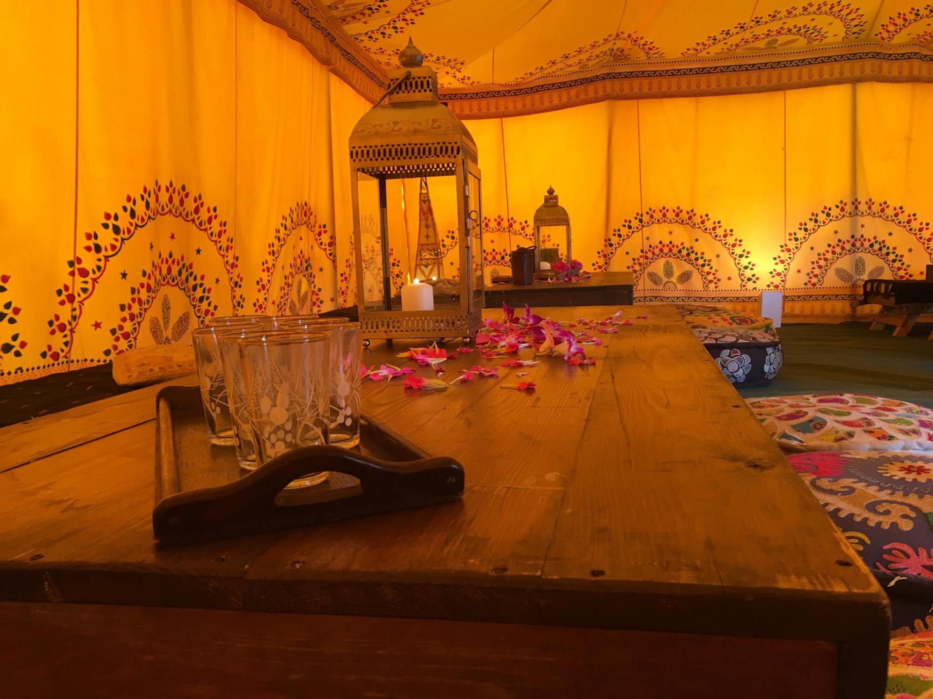 Interior of a yellow tent with a candlelit lantern and a tray of glasses and rose petals on a wooden table.