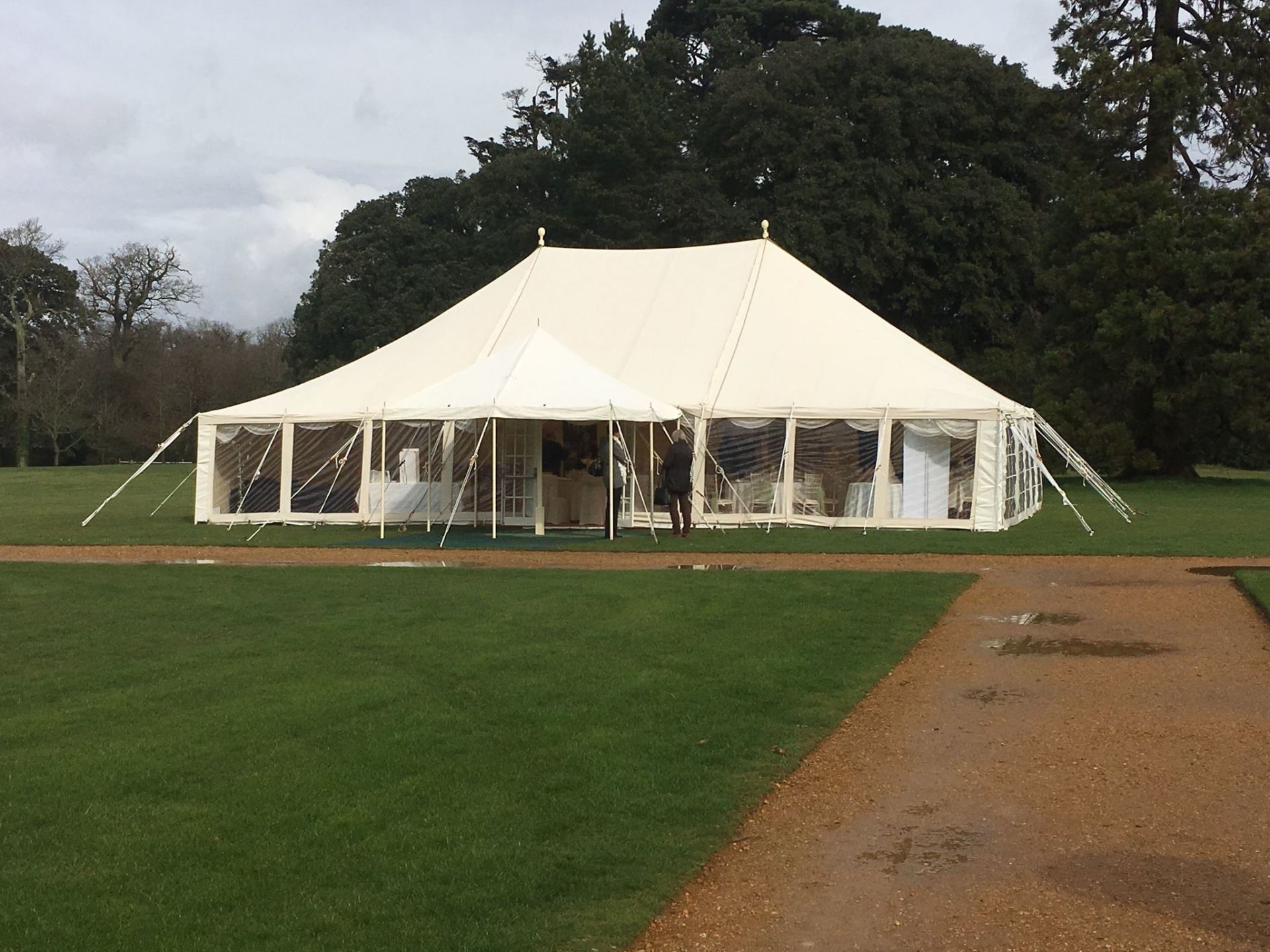White marquee tent with clear windows, set on a green lawn, near a paved path and trees.