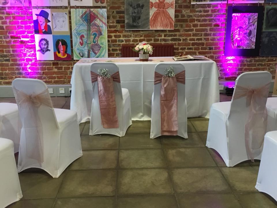 Wedding reception setup: chairs with pink sashes face a table covered in a white cloth, with pink up-lighting against a brick wall.