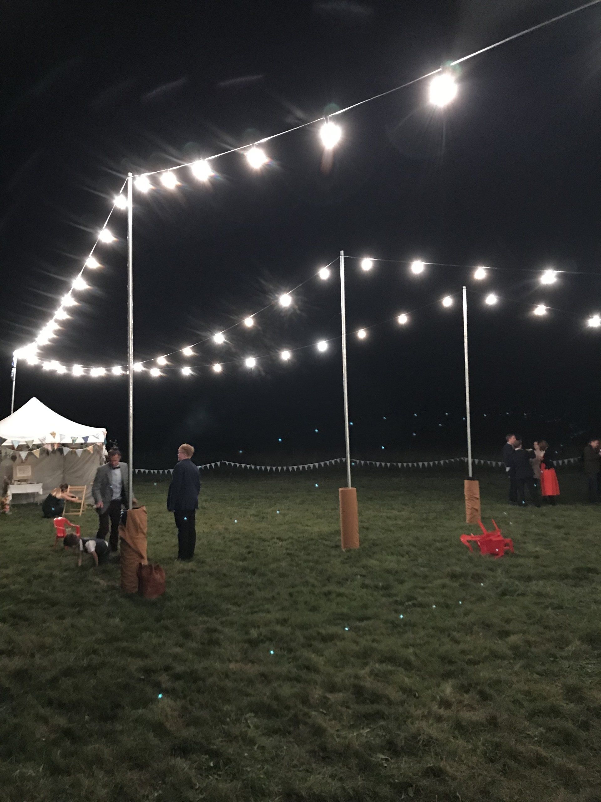 String lights illuminate a grassy field at night. Three poles support the lights, with people near the center. A white tent is on the left.