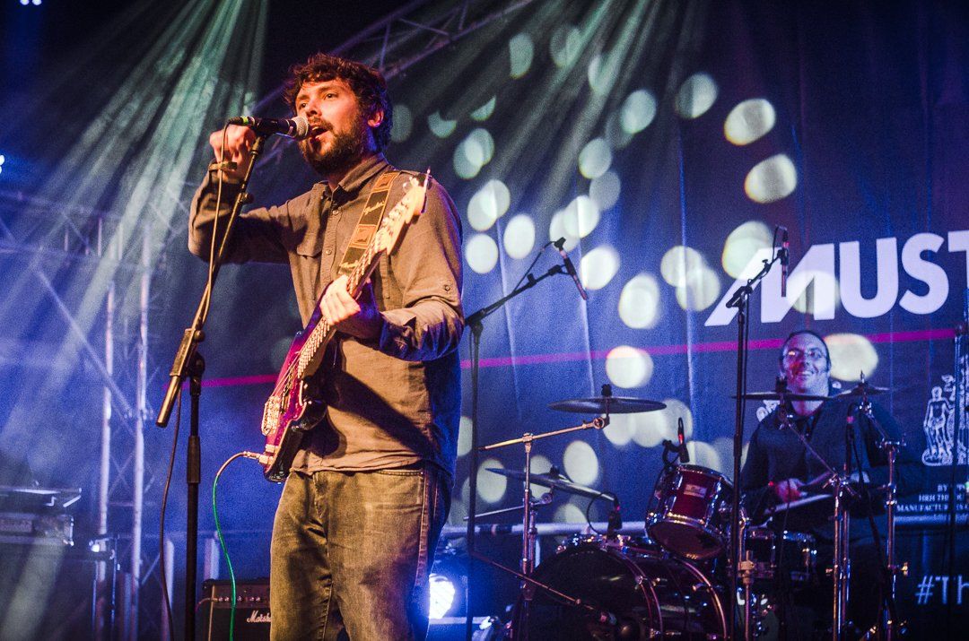 Man singing and playing guitar onstage with a drummer in a venue with stage lights.