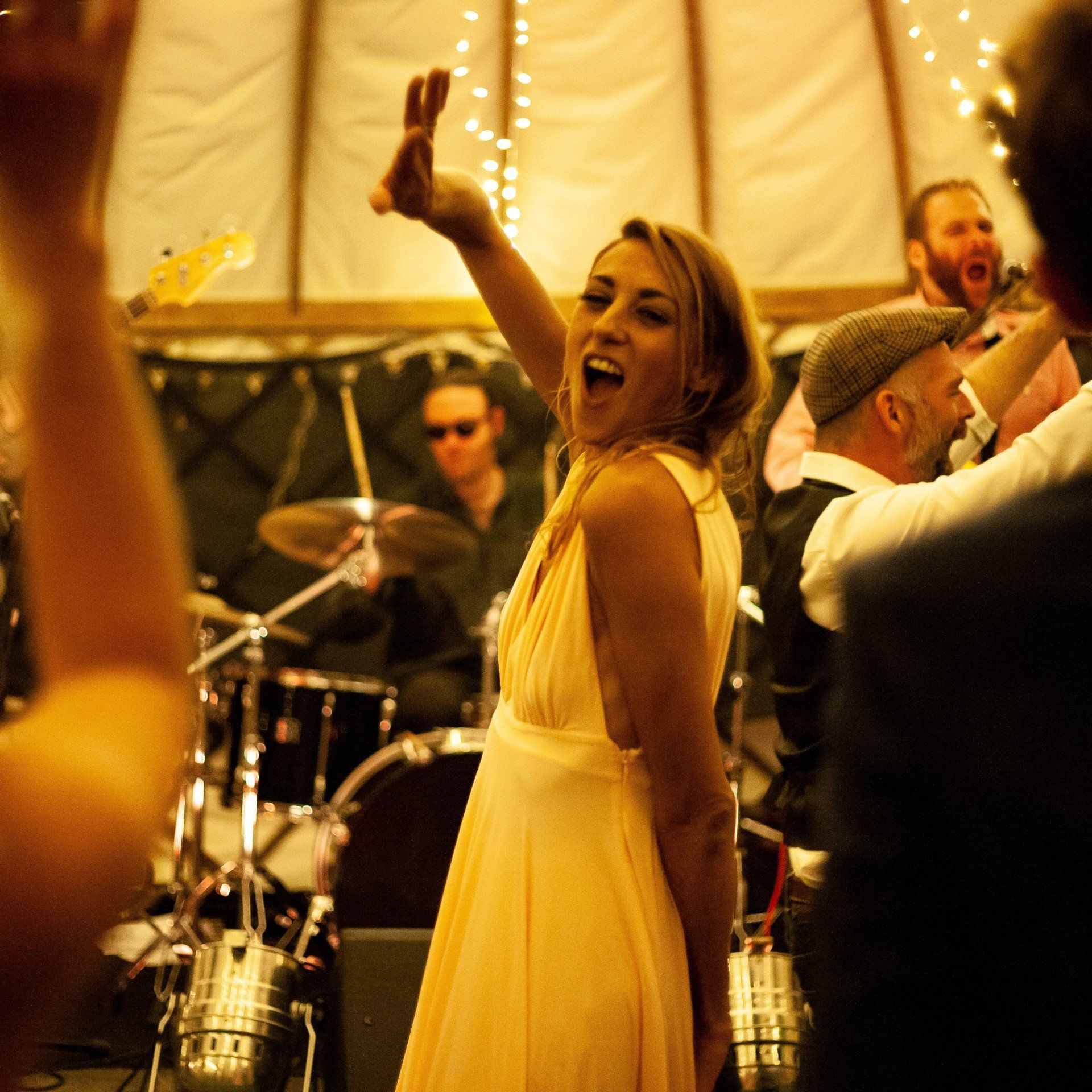 Woman in yellow dress singing on stage with band, raising arm. Lit tent setting.