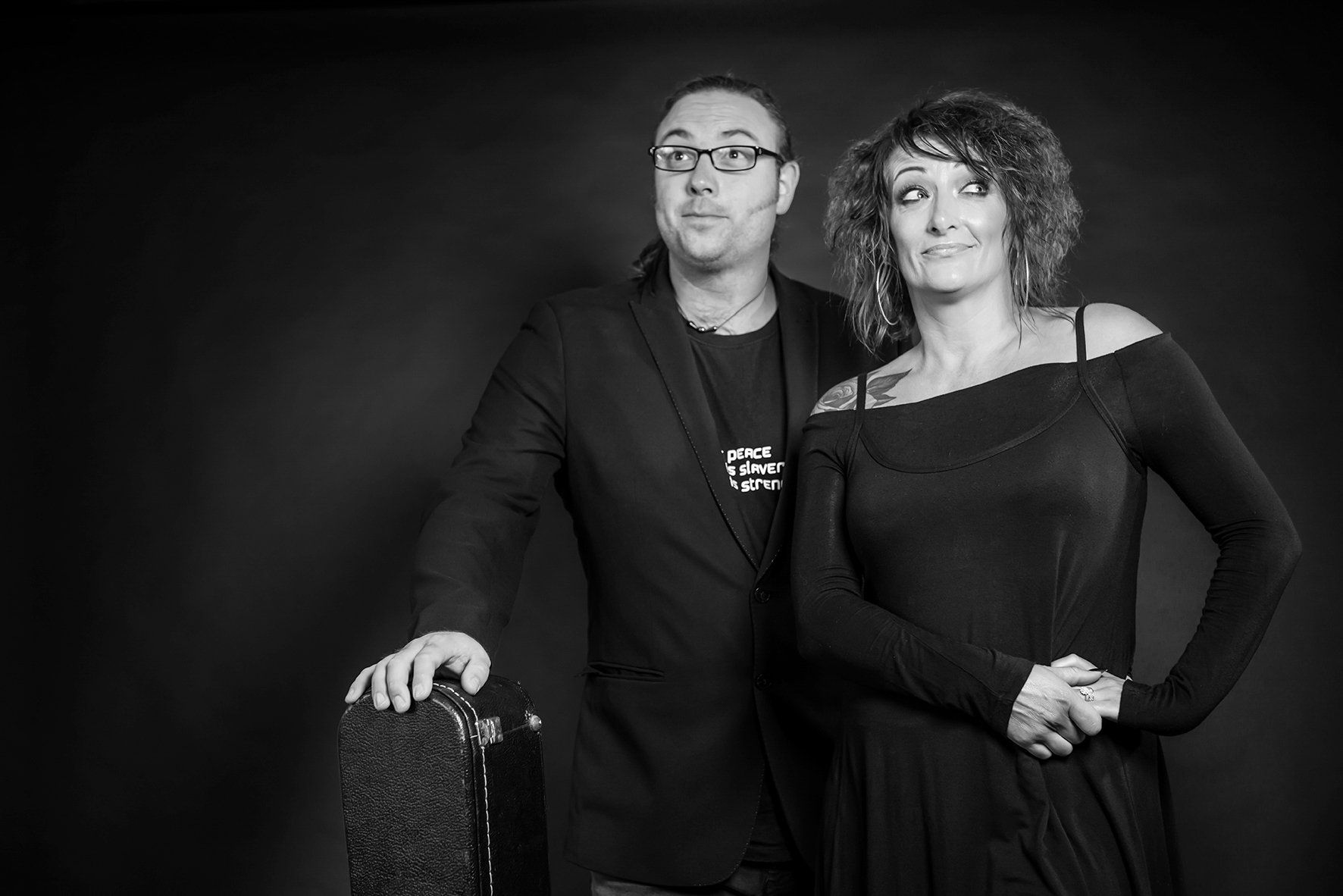 Man in glasses and black jacket, leans on chair next to woman in off-the-shoulder dress, both smiling, black and white photo.