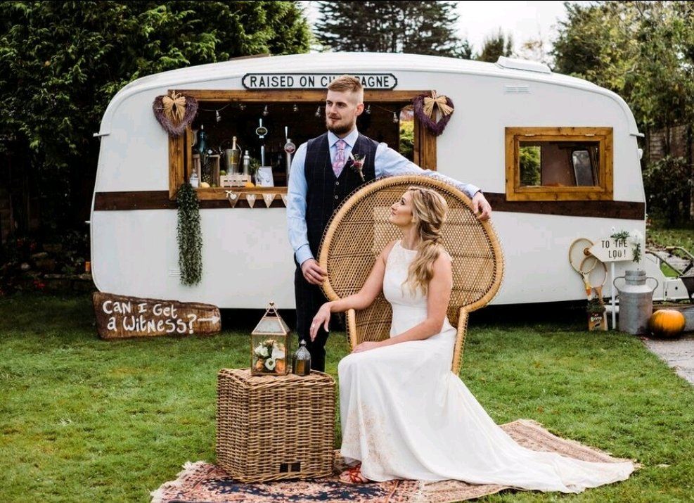 Newlyweds pose next to a vintage bar caravan in a garden, she in a white dress, he in a vest.