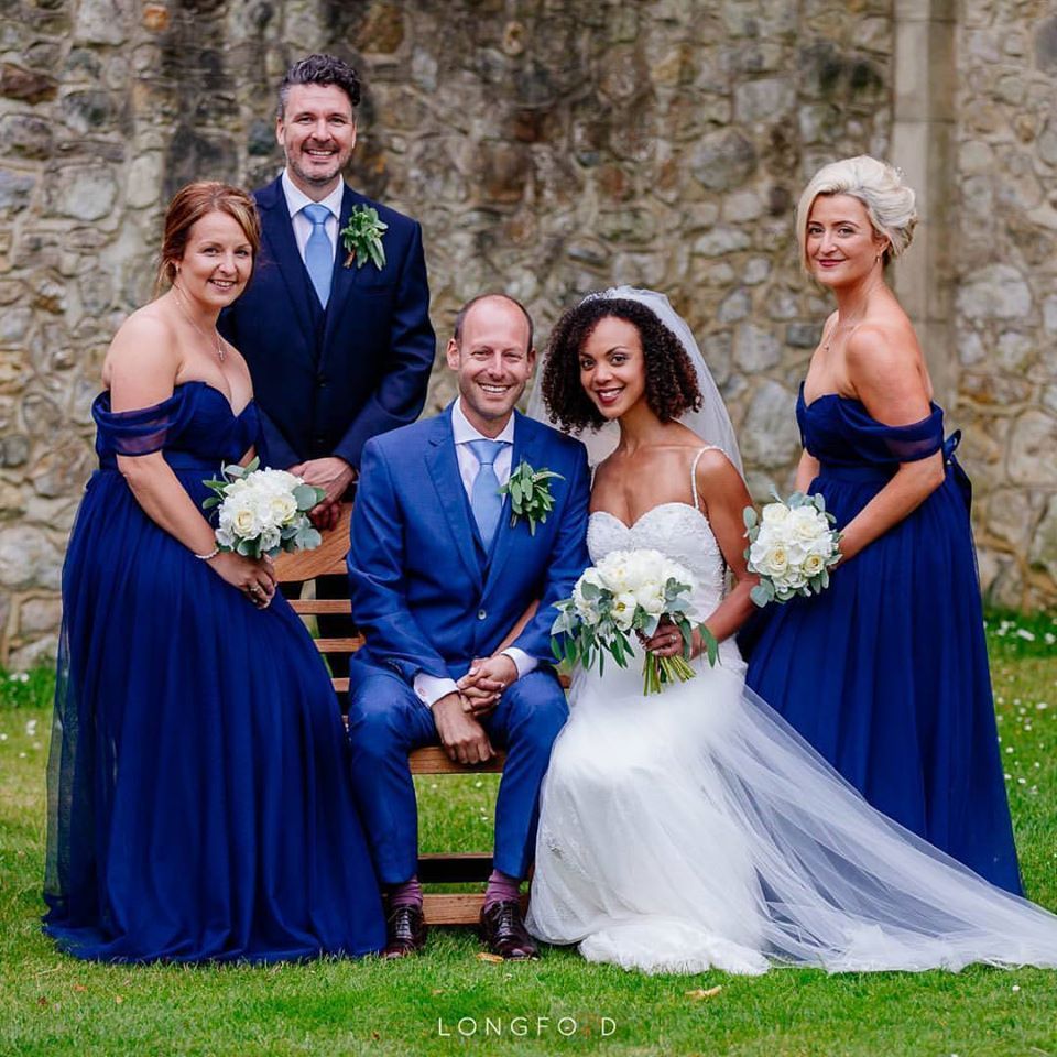 Wedding party posing in front of a stone wall. Bride and groom seated, flanked by bridesmaids and a groomsman, all wearing formal attire.