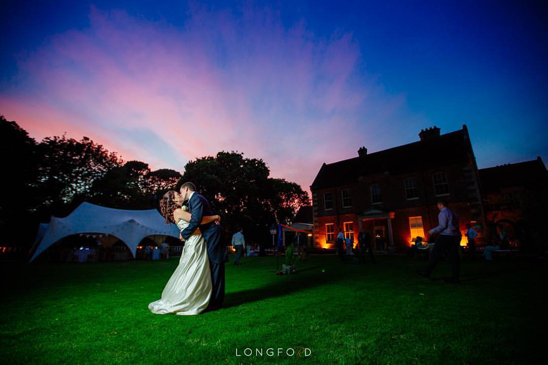 Couple embracing on a lawn in front of a house, twilight sky overhead.