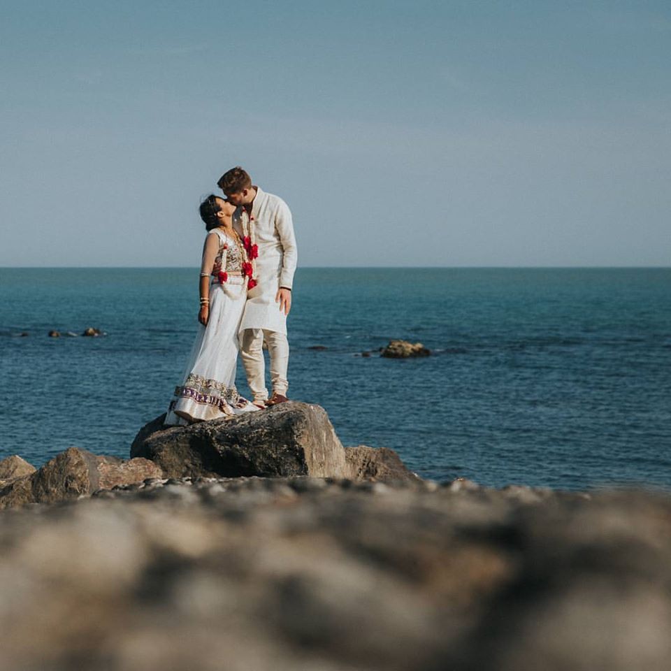 Couple kissing on a rock by the ocean. She wears a white outfit with red flowers. He wears a cream suit.